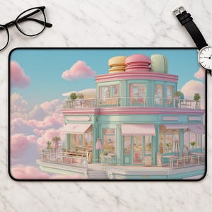 May include: A rectangular desk mat featuring a whimsical two-storey building with mint green walls and pink accents, topped with large yellow, pink, and green macaron sculptures. The structure is surrounded by soft pink clouds in a blue sky.