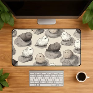 May include: A rectangular desk mat with a repeating pattern of cartoon seals on gray rocks. The seals are in various shades of grey and white against a beige background. The mat is surrounded by a wooden desk, a keyboard, and a coffee cup.