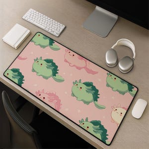 May include: A rectangular desk mat featuring a repeating pattern of cartoon axolotls in shades of light green and soft pink against a solid pink background, accented with small white star shapes across the entire surface of the mat.