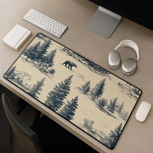 May include: A rectangular desk mat featuring a cream-coloured background with a dark blue illustrative print of a forest landscape. The design includes tall pine trees, a walking bear, a deer, and small birds flying over a mountainous terrain.