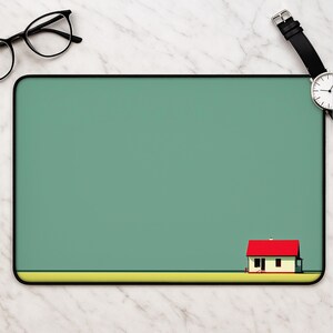 May include: A rectangular desk mat featuring a solid sage green background with a thin yellow horizontal stripe at the bottom. A small, simplified house with a bright red roof and cream-colored walls is positioned in the bottom right corner.