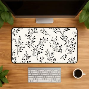 May include: A rectangular desk mat featuring a repeating pattern of black botanical branches with small leaves on a cream-colored background. The mat has a thin black border and is positioned horizontally on a wooden surface below a computer monitor.