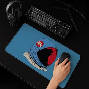 Può includere: Un tappetino da scrivania rettangolare con sfondo blu caratterizzato da un'illustrazione grafica di un casco da astronauta. Il casco è blu con una visiera rossa e una toppa circolare rossa sul lato contenente il testo bianco MTL. Il tappetino ha un bordo nero.