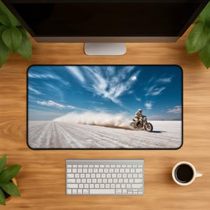 Puede incluir: Alfombrilla de escritorio rectangular con una motocicleta corriendo por un salar blanco bajo un cielo azul con nubes. La motocicleta levanta una nube de polvo. Se ven un teclado y una taza de café.