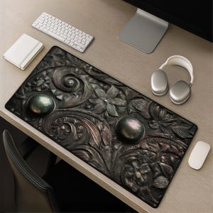 May include: A dark, intricately carved desk mat with floral and swirling patterns. Two large, iridescent, dark green pearls are set on the mat. A white keyboard, mouse, headphones, and notepad are also visible.