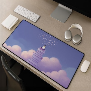 May include: A rectangular mouse mat with a charming illustration of a penguin on a pile of books, set against a backdrop of clouds and hearts. The mat has a black border and a smooth surface. A white keyboard, mouse, headphones, and notepad are also visible.