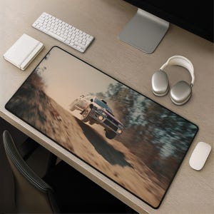 May include: A rectangular desk mat featuring a high-speed action photograph of a rally car jumping over a dirt mound. The car is captured mid-air against a blurred background of trees and a dusty track, with motion blur emphasizing the speed.