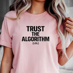 May include: A light pink t-shirt with the text "TRUST THE ALGORITHM (LOL)" printed in black. The shirt has short sleeves and is paired with light blue denim shorts. The overall style is casual and relaxed.
