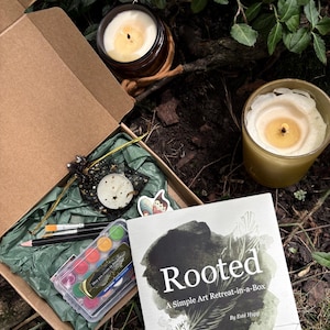 May include: An open cardboard box with art supplies, a book, and two lit candles. The box contains watercolor paints, paintbrushes, pencils, and a small bag. The book cover reads "Rooted: A Simple Art Retreat-in-a-Box" by Esté Hupp.