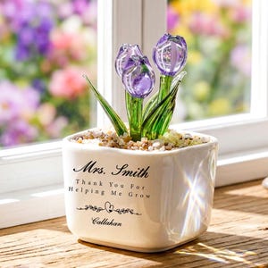 Puede incluir: Una maceta de cerámica blanca con tres tulipanes de cristal morado. La maceta tiene el texto "Mrs. Smith Thank You For Helping Me Grow Callahan" en escritura negra. La maceta está sobre un alféizar de madera con un fondo floral borroso.