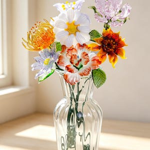 May include: A clear glass vase with vertical fluting containing a bouquet of handcrafted glass flowers. The arrangement features a white daisy, an orange spider mum, a purple star-shaped flower, a pink blossom, and a red and white variegated flower.