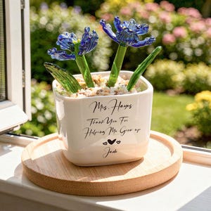 May include: A white square ceramic planter filled with small stones and two blue glass flowers with green stems. The planter features black text that reads: Mrs. Hayes, Thank You For Helping Me Grow up, followed by two small hearts and the name Jade.