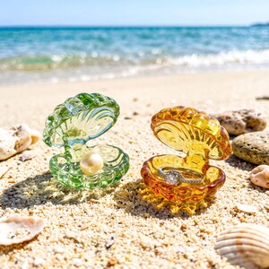 May include: Two open seashell-shaped trinket boxes made of translucent glass. One box is green and contains a single white pearl. The other box is amber-colored and holds a silver-toned ring with a central stone and small pavé accents.