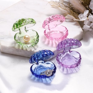 May include: Four small, open, clam-shaped glass trinket boxes in green, pink, purple, and blue. The green box contains a single cream-colored pearl, and the blue box holds a silver-toned ring with a clear stone setting. Each box has a ribbed exterior.