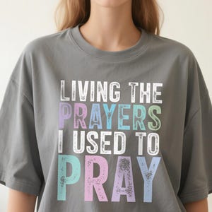 May include: Gray t-shirt with the phrase "LIVING THE PRAYERS I USED TO PRAY" in a distressed, multi-colored font. The text is in white, purple, blue, and green. The shirt has short sleeves and a crew neck.