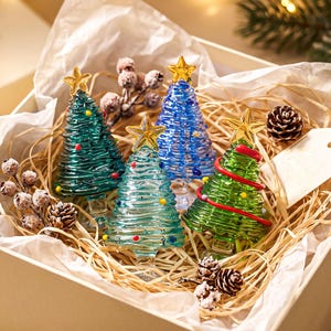 May include: Four glass Christmas tree figurines in dark green, light blue, royal blue, and lime green, each topped with a gold star. The trees feature horizontal ribbed textures and small colourful dots, nestled in a box with straw and pinecones.