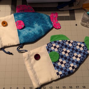 May include: Two handmade fish-shaped pouches. One has a blue tie-dye pattern, pink accents, and a wooden button. The other features a blue and white checkered pattern with green accents and a purple button. Both have white fabric and drawstring closures.