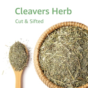 May include: A wooden spoon filled with cut and sifted cleavers herb next to a woven basket filled with the same herb. The text "Cleavers Herb" and "Cut & Sifted" are in green font above the basket and spoon.