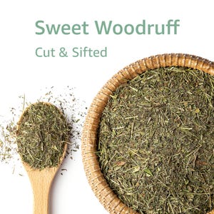 May include: A top-down shot shows Sweet Woodruff, cut and sifted, in a woven basket and a wooden spoon. The dried herb is green and brown, with the text "Sweet Woodruff" and "Cut & Sifted" in green above the basket.