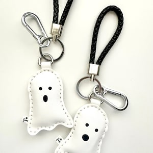 May include: Two white ghost-shaped keyrings with black eyes and mouths, stitched with white thread. Each keyring has a black braided leather strap, a silver-tone metal clasp, and a small silver ring.
