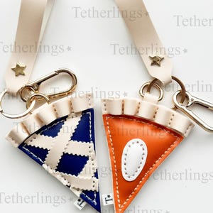 May include: Two pie slice keychains with cream-colored straps and gold-tone hardware. One keychain is blue and cream, the other is orange and white. Each has a star accent and a lobster clasp.