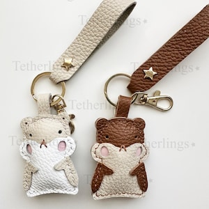 May include: Two leather keychains, one beige and one brown, each featuring a bear-shaped charm. The charms have white and brown details with pink accents. Each keychain has a matching strap with a gold star and a keyring.