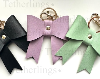 May include: Three leather bow keychains in black, lavender, and mint green. Each bow is attached to a gold-colored keyring with a lobster clasp. A gold-colored stud secures the center of each bow.