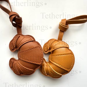 May include: Two leather croissant-shaped keyrings in shades of brown and tan. Each keyring has a loop for attachment and is stitched with a contrasting thread. The keyrings are designed to resemble a croissant pastry.