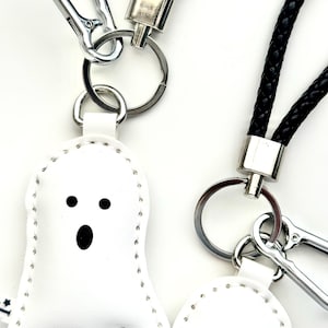 May include: White ghost-shaped keyrings with black facial features and silver key rings. The keyrings have a stitched outline and are attached to black braided wrist straps and silver carabiner clips. The background is white.