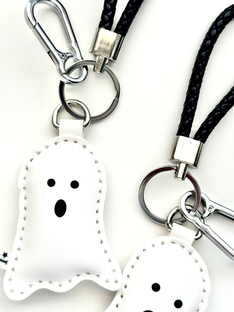 May include: White ghost-shaped keyrings with black facial features and silver key rings. The keyrings have a stitched outline and are attached to black braided wrist straps and silver carabiner clips. The background is white.