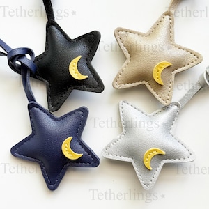 Puede incluir: Cuatro colgantes en forma de estrella con lazos, cada uno con una pequeña aplicación de luna creciente amarilla. Los colgantes son de color negro, azul marino, beige y plateado, con costuras visibles en los bordes. El texto Tetherlings es visible.