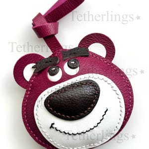 May include: A magenta leather keychain shaped like a bear's face, featuring white accents around the eyes and mouth, and a dark brown nose. The keychain has a loop at the top for attachment. The text "Tetherlings" is visible in the background.