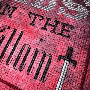 K&ouml;nnte beinhalten: Kreuzstich-Design mit den Worten "WORDS ON THE SILENT" in schwarzem und wei&szlig;em Garn auf rotem und rosa Hintergrund. Unter dem Text ist ein detailliertes Schwert gestickt, das dem St&uuml;ck ein dekoratives Element hinzuf&uuml;gt.