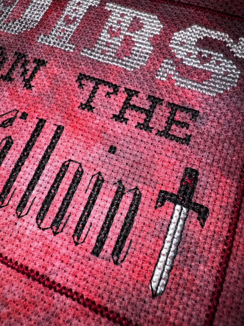 K&ouml;nnte beinhalten: Kreuzstich-Design mit den Worten "WORDS ON THE SILENT" in schwarzem und wei&szlig;em Garn auf rotem und rosa Hintergrund. Unter dem Text ist ein detailliertes Schwert gestickt, das dem St&uuml;ck ein dekoratives Element hinzuf&uuml;gt.