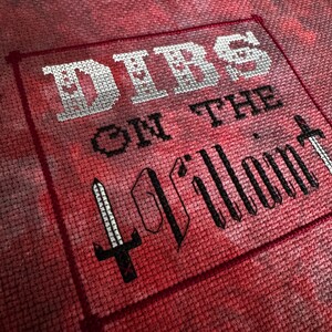 K&ouml;nnte beinhalten: Kreuzstich-Textil mit den Worten "Dibs on the Villain" in Wei&szlig; und Schwarz. Der Text ist von einem roten Rahmen umgeben und zeigt eine Schwertgrafik. Der Hintergrund hat einen roten und rosa Batik-Effekt.