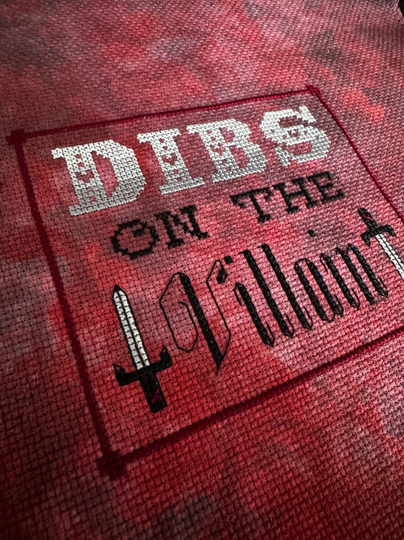 K&ouml;nnte beinhalten: Kreuzstich-Textil mit den Worten "Dibs on the Villain" in Wei&szlig; und Schwarz. Der Text ist von einem roten Rahmen umgeben und zeigt eine Schwertgrafik. Der Hintergrund hat einen roten und rosa Batik-Effekt.