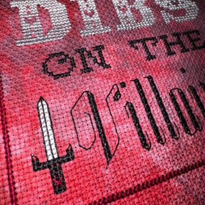 K&ouml;nnte beinhalten: Nahaufnahme eines Kreuzstichdesigns auf rotem Stoff. Der Text lautet "DIBS ON THE PILLOW" in wei&szlig;em und schwarzem Garn. Auf der linken Seite ist ein schwarzes Schwert gestickt. Der Stoff hat ein strukturiertes Aussehen.