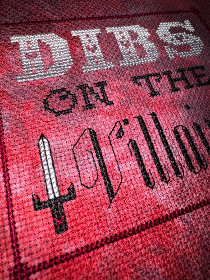 K&ouml;nnte beinhalten: Nahaufnahme eines Kreuzstichdesigns auf rotem Stoff. Der Text lautet "DIBS ON THE PILLOW" in wei&szlig;em und schwarzem Garn. Auf der linken Seite ist ein schwarzes Schwert gestickt. Der Stoff hat ein strukturiertes Aussehen.