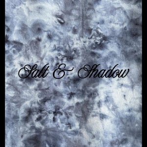 May include: A textile featuring a grey and white tie-dye design. The words "Salt & Shadow" are written in cursive across the bottom of the fabric.