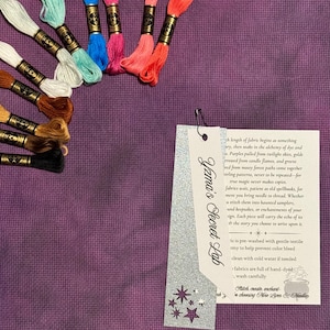 May include: A collection of colorful embroidery floss skeins in shades of white, teal, blue, pink, orange, brown, and black, arranged on a purple fabric. A tag with the text "Yzma's Secret Lab" is attached with a silver paperclip.