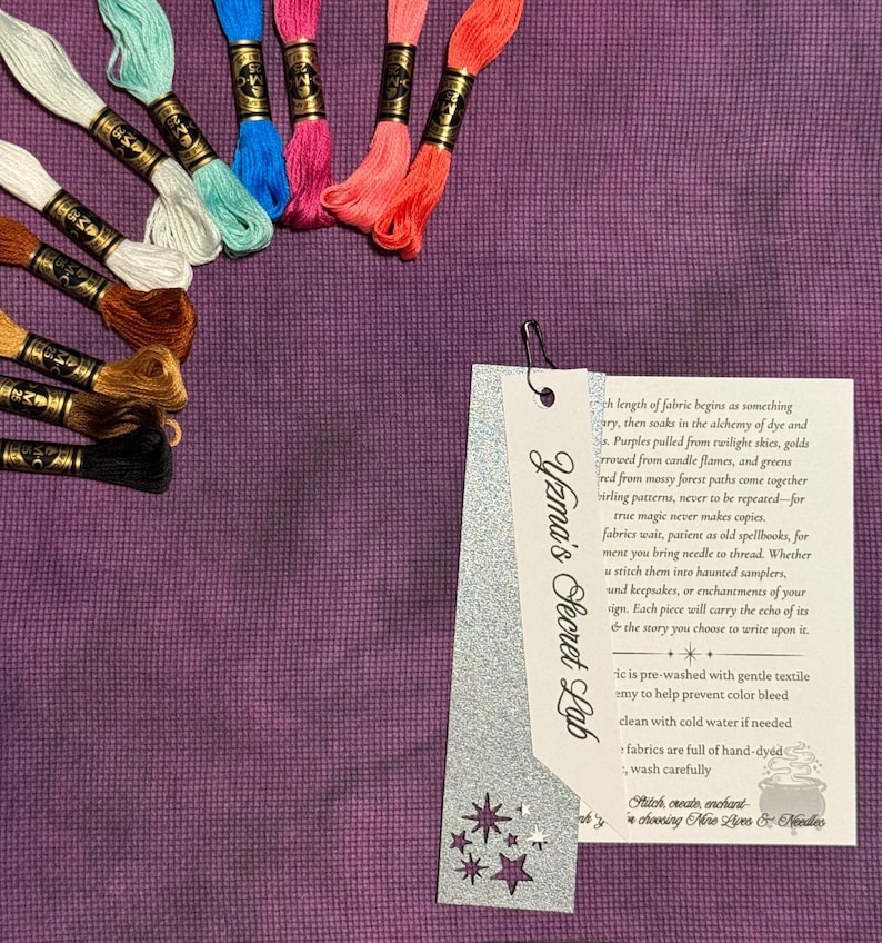 May include: A collection of colorful embroidery floss skeins in shades of white, teal, blue, pink, orange, brown, and black, arranged on a purple fabric. A tag with the text "Yzma's Secret Lab" is attached with a silver paperclip.