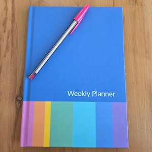 May include: A blue weekly planner with a colorful striped design at the bottom and the words "Weekly Planner" in white. A pink and silver pen rests on top of the planner.