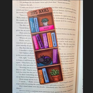 May include: A colorful bookmark featuring a bookshelf design with the text "2025 BOOKS." The bookmark displays various book titles, a sleeping dragon, and potted plants. It is inserted into the pages of a book.