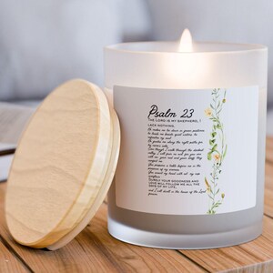 Puede incluir: Una vela de vidrio esmerilado encendida con una tapa de madera está sobre una mesa de madera. La vela tiene una etiqueta con el texto "Salmo 23" e ilustraciones florales. La vela está junto a su tapa de madera.