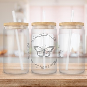 May include: Three clear glass drinking jars with wooden lids and clear straws. The center jar features a butterfly illustration and the text "...being beautiful in its time...He has made...3:11". The jars are on a wooden surface.