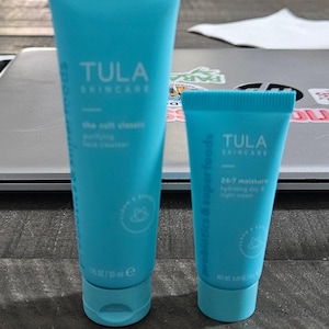 Może przedstawiać: Dwie turkusowe tubki produktów TULA Skincare. Większa tubka jest oznaczona jako "the cult classic purifying face cleanser" i zawiera 50 ml. Mniejsza tubka jest oznaczona jako "24-7 moisture hydrating day & night cream" i zawiera 17 g.