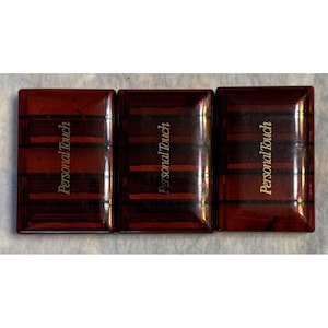 May include: Three rectangular, dark red cosmetic compacts with the words "Personal Touch" in gold lettering. The compacts are connected and have a glossy finish. The design includes vertical and horizontal lines.