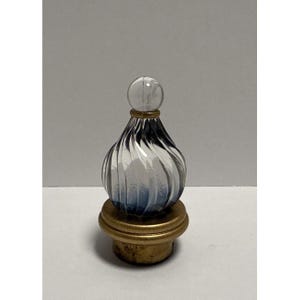May include: A decorative bottle stopper featuring a clear glass orb top and a teardrop-shaped glass body with blue and white swirling patterns. The base is constructed from a gold-toned metal ring and a cork stopper for a secure fit in bottles.