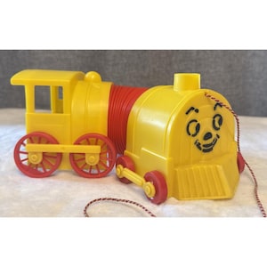 May include: A yellow toy train with red wheels and a red accordion-style connector. The front of the train has a smiling face. A string is attached for pulling the toy.