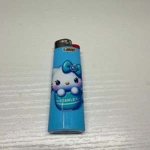 以下が含まれることがあります： ハローキティのデザインが施された青いBICライター。ライターには赤い点火ボタンと、名前「Stanley」がプリントされた白と青のハローキティのグラフィックがあります。